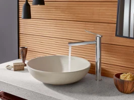 Une salle de bains moderne avec une vasque à poser ronde Loop & Friends, de l’eau s’écoule d’un robinet minimaliste, des panneaux muraux en bois et divers articles de toilette sur un plan de toilette gris.