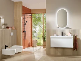 A stylish beige bathroom featuring the Skyla bidet, toilet, and washbasin, complemented by a terracotta accent wall in the shower area. The More To See Lite illuminated mirror adds modern functionality and soft, ambient lighting.