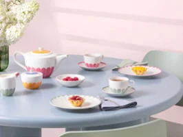 A pastel-colored tea set with cups, saucers, plates with colorful floral patterns from the Fleur collection, and small desserts on a light blue round table.