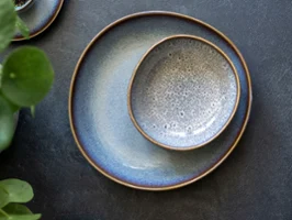 Deux assiettes Lave avec un émail beige à effet sur une surface structurée sombre, accompagnées d’une plante verte.