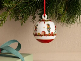 Annual Chistmas edition 2026 ball A white Christmas tree ornament featuring a festive scene by Villeroy & Boch hangs from a red string attached to a fir branch above a wrapped gift.