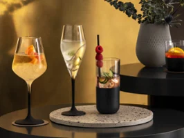 Black Manufacture glasses with cocktails A selection of four elegant cocktails in different glasses, garnished with fruits and herbs, arranged on a table with a plant in the background.