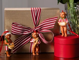 Three small bear figurines from Villeroy & Boch stand on a table next to a wrapped gift with a striped ribbon and a round red box.