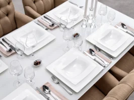 A beautiful dining table setting with square white crockery from the Villeroy & Boch Modern Grace brand, silver cutlery, wine glasses and beige napkins. Beige upholstered chairs surround the table. There are three candlesticks in the centre.