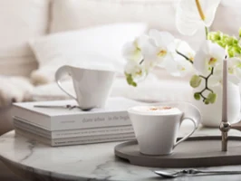 Zwei Cappucchino-Tassen von Villeroy & Boch NewWave, einem eleganten Tablett, dekorativen Büchern und einer Kerze. Weiße Blumen verleihen dem Hintergrund eine charmante Note.