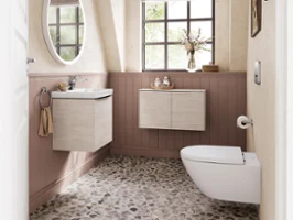 A bathroom with a window, pebble floor, wall-mounted washbasin and a toilet. Above the washbasin there is a round mirror, on the lower part of the walls wood panelling, with framed pictures higher up on the walls.
