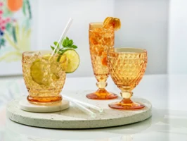 Boston Saffron colored crystal glasses with refreshing summer drinks Three Villeroy & Boch Boston crystal glasses in vibrant saffron orange, including a tumbler with lime and mint, a champagne flute with physalis, and a wine goblet, arranged on a round terrazzo board on a white table.