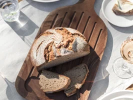 Ein Brotlaib mit zwei Scheiben ruht auf einem Artesano Holzbrett von Villeroy & Boch. Auf dem Tisch stehen Teller, Servietten, ein Glas Wasser, eine Schale Oliven und ein Glas Wein.
