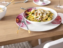 Ein Rose Garden Pastateller mit Nudeln, serviert auf einem Holztisch, dazu Ella Besteck mit goldenen Akzenten und ein Glas Wasser.