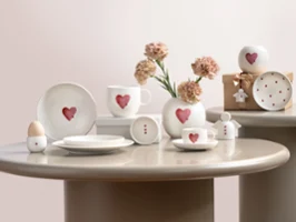 Vaisselle en céramique de la marque With Love avec motifs en forme de cœur rouge, disposée sur des tables rondes avec des fleurs et un coffret cadeau.