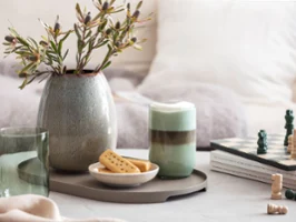  Like Sage Glas mit Kaffee, einem Schachbrett, Keksen und einer Lave Vase mit Pampasgras auf einer Tischplatte.