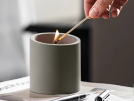 A hand lighting a Villeroy & Boch NewMoon candle with a match, on a table with paper and a pen.