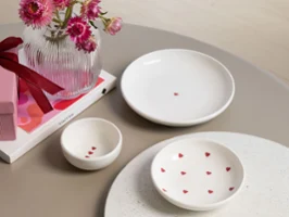 Drei weiße Keramikteller mit kleinen roten Herzmotiven von der Kollektion with love stehen auf einem Tisch neben einer Vase mit rosa Blumen und einer Geschenkbox.