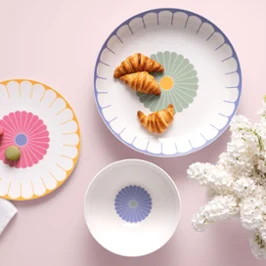 Three Villeroy & Boch Fleur plates with croissants and macarons next to white flowers on a light pink background.