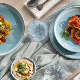 A table set with pasta, salad, hummus, olives and utensils on blue plates from the Perlemor Aqua collection and a light-coloured tablecloth.