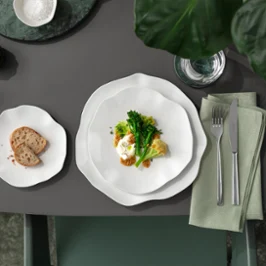 A Pura plate with vegetables and egg, a side plate with bread and cutlery on a napkin on a grey table.