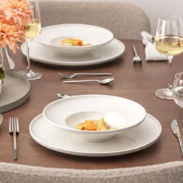 A neatly laid dining table from the Afina collection with two plates of gourmet food, wine glasses, water glasses, silver cutlery, cloth napkins and a vase of peach blossoms on a wooden surface.
