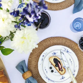 Table setting with a slice of cake and blueberries on an Old Luxembourg plate, surrounded by a woven placemat, cutlery and a blue napkin. A vase of white and purple flowers can be seen at the side.