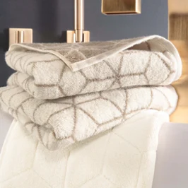 Une pile de serviettes « Carré » beiges pliées à motifs géométriques de Villeroy & Boch, placées à côté d’un robinet doré.