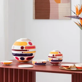 Colorful, multi-piece tableware sets from la petite boule miami, circular design objects in large and small sizes, stand as decoration next to a vase with abstract patterns and orange flowers on a red table.
