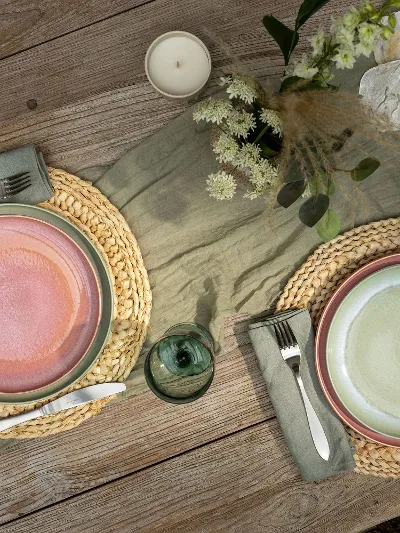 Two Perlemor place settings on a wooden table with round woven placemats, cutlery, a candle, flowers and green plants.