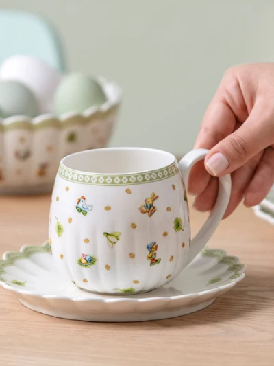 A hand holds a Easter Delight teacup on a saucer, with a bowl of eggs and plates in the background on a wooden table.