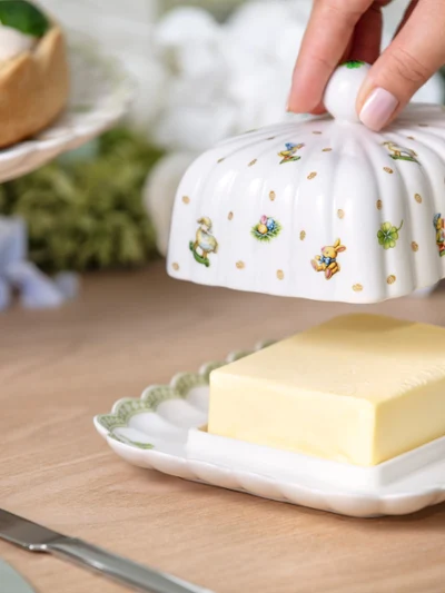 A hand lifts the lid of a decorative butter dish with an Easter pattern from Easter Delight containing a rectangular block of butter on a wooden table.