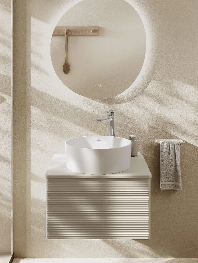 Un lavabo da appoggio bianco rotondo su un mobile sottolavabo in legno con rubinetteria cromata e specchio dalla forma organica.