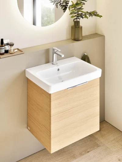 White Architectura washbasin with a light-coloured wooden cabinet and an elegant tap Modern bathroom with a rectangular white washbasin, an elegant tap and a light-coloured wooden cabinet. Above the washbasin is a round mirror, on the shelf there are toiletries and a small vase with a plant.