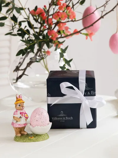 A Villeroy & Boch Bunny Tales egg cup on a white table next to a gift box wrapped with a bow. In the background is a vase with twigs and pink flowers.