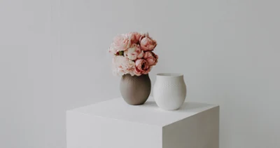 A brown vase with pink flowers and a white ceramic vase stand on a white cube against a plain light-coloured background.