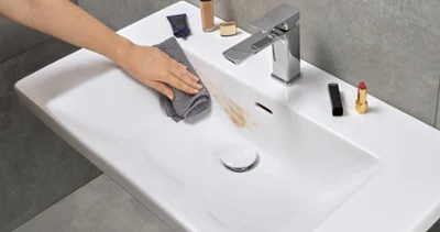 Czyszczenie zabrudzonej umywalki Ręka czyszcząca szarą ściereczką umywalkę w łazience i usuwająca plamę z produktu do makijażu. Na krawędzi umywalki znajdują się różne produkty do makijażu.