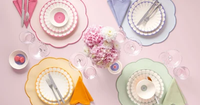 Buntes Tablesetting mit farbigen Fleur Tellern, Schalen und Platzsets Vier Platzsets mit farbigen Tellern und Schalen aus der Fleur Kollektion, farbigen Servietten und Besteck sind um einen rosa Blumenschmuck angeordnet.