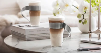 NewWave - Kaffee elegant genießen Zwei geschichtete Lattes in modernen NewWave Glasbechern mitg eschwungenen Metallgriffen stehen auf einem Marmortisch mit Büchern, Blumen und Kerzen.