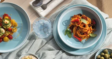 A table set with pasta, salad, hummus, olives and utensils on blue plates from the Perlemor Aqua collection and a light-coloured tablecloth.