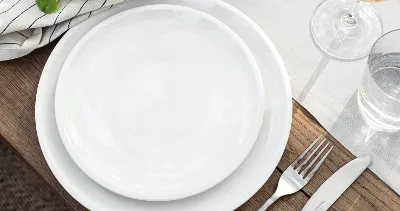 An Artesano wooden table set from Villeroy & Boch with white plates, a fork, a knife, a glass of water, a wine glass, and a striped napkin with green leaves.