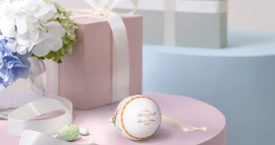Ornement de Pâques annuel blanc doré sur une table rose Une boîte cadeau rose pastel ornée d'un nœud blanc est posée sur une table rose, à côté d'une décoration de Pâques annuelle blanche et dorée, d'un nœud, de bonbons vert clair et d'un vase contenant des fleurs blanches et bleues. Une boîte cadeau bleue est posée à l'arrière-plan.