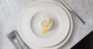 A small portion of food on a white Manufacture plate A small portion of food is arranged on a white Manufacture plate with a knife, fork, and napkin on a marble surface.