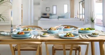 A wooden dining table for four people with blue Perlemor Aqua plates in a bright, modern living area.