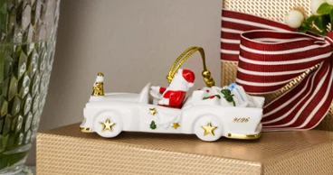 Cadillac 2026 gift A ceramic Christmas car with presents is sitting on a gift wrapped in gold, next to a vase of red and white flowers.
