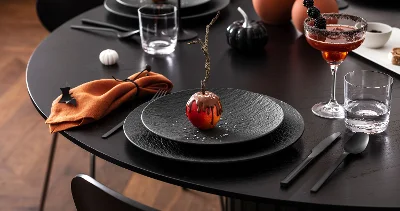 Un tavolo apparecchiato in nero con piatti neri, posate nere, tovaglioli arancioni, decorazioni e bevande a tema Halloween.