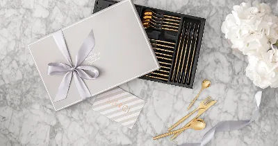 Gift box with gold MetroChic cutlery and a bow, a thank you card and white flowers on a marble surface.