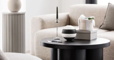 A round black coffee table with the Villeroy & Boch La petite Boule A round black coffee table with the Villeroy & Boch La petite Boule, a green candle on a silver holder and a gift box on top of two books. Beige sofa and a grey plinth with a vase in the background.