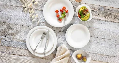 A rustic table setting with Essential White and New Cottage plates, fork and knife, a beige napkin and bowls of cherry tomatoes, avocado slices, and figs. On the top right is a glass of rosé wine.