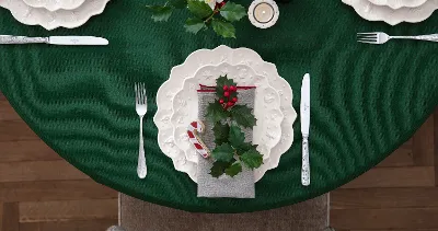 A Christmas table setting with a green tablecloth, white plates decorated with holly from the Villeroy & Boch Toy's Delight Royal Classic collection, a candy cane, a sprig of holly on the napkin and silver cutlery.