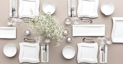 The Villeroy & Boch NewWave table set consisting of square white plates, bowls, cutlery, glasses and a centrepiece with white flowers on a beige tablecloth for four people.