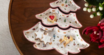Toy's Delight 3-piece tree-shaped snack bowl set Overhead view of a 3-piece Christmas tree-shaped porcelain snack bowl set from Toy's Delight collection on a dark wooden table, filled with festive star cookies and a heart-shaped biscuit, next to a red gift box with a green-and-white striped ribbon and deep red flowers