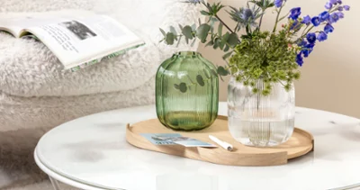 Ein runder Couchtisch mit einer Villeroy & Boch Fleur Vase, die blaue und grüne Blumen hält. Ein runder Couchtisch mit einer Villeroy & Boch Fleur Vase, die blaue und grüne Blumen hält. Im Hintergrund liegt eine aufgeschlagene Zeitschrift auf einem weißen, flauschigen Stuhl.