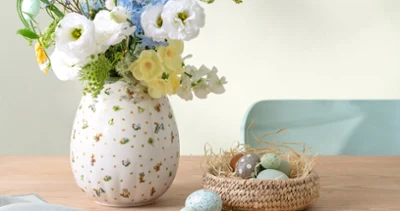 A Easter Delight vase with white and blue flowers sits beside a small nest filled with pastel eggs on a wooden table.