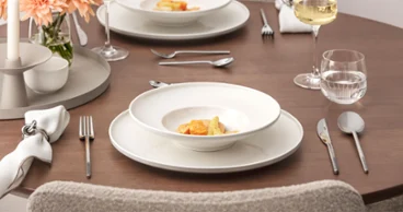 Ein ordentlich gedeckter Esstisch der Afina Kollektion mit zwei Tellern mit Gourmetgerichten, Weingläsern, Wassergläsern, Silberbesteck, Stoffservietten und einer Vase mit Pfirsichblüten auf einer Holzfläche.